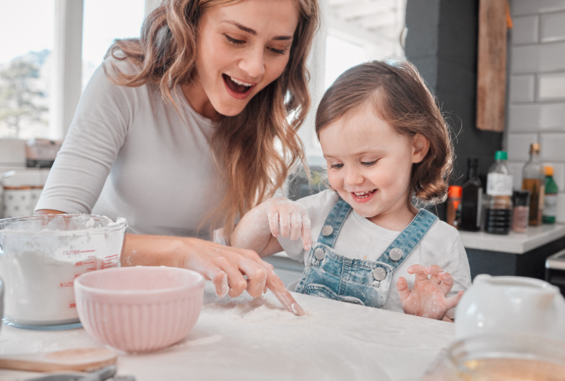 Picture shows a short-term nanny cooking with a smiling child. Kensington nanny services also provide short-term care.