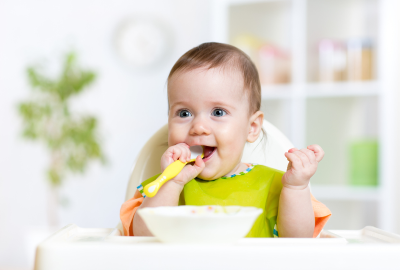 happy baby is sitting in a high chair eating lunch being served by a babysitter in south florida. In Kensington Nanny services page, this is under nanny agency pricing.