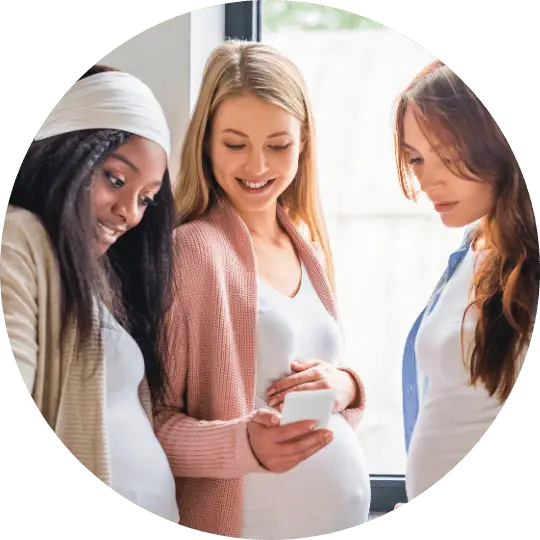3 pregnant women talking. 1 woman is showing the Kensington Nanny website on her phone. Giving Kensington Nanny a great nanny agency review.