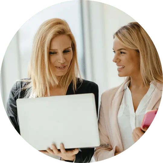 2 women are discussing Kensington Nanny. One woman is holding a laptop and showing the other Kensington's website. She is giving Kensington Nanny a great nanny agency review.